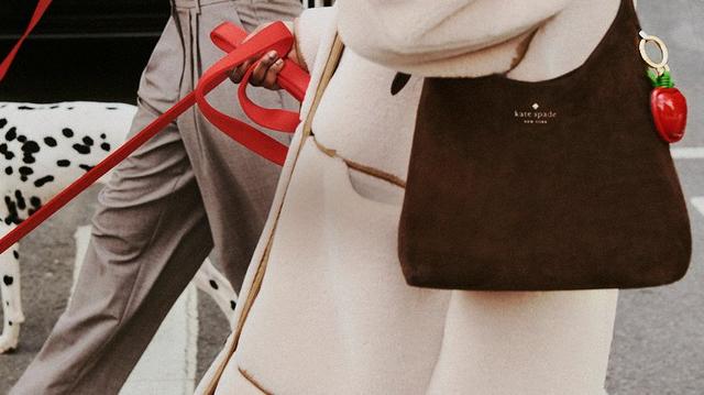 Kate spade new york brown shoulder bag with strawberry charm, held by person walking dalmatian dog.
