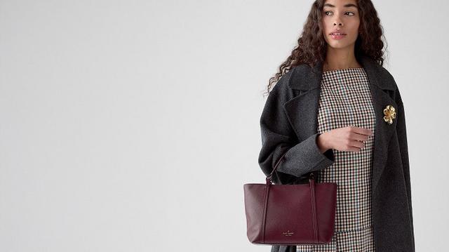 Person wearing a gray coat and checkered dress holding a burgundy leather handbag with a gold brooch.