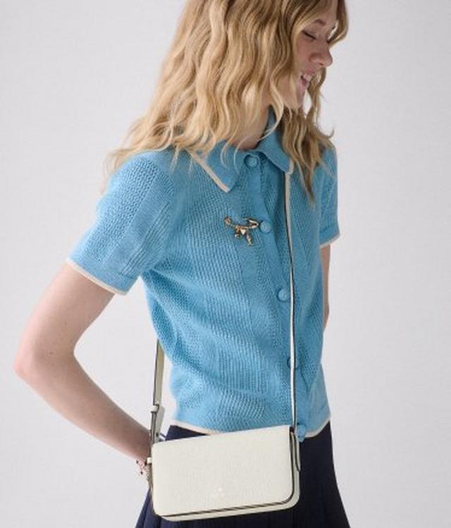 Woman wearing blue knit polo shirt with decorative pin, carrying white crossbody bag.