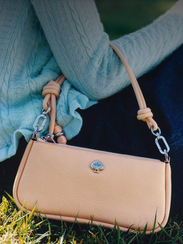 Person holding a peach-colored Kate Spade leather crossbody bag while sitting on grass.