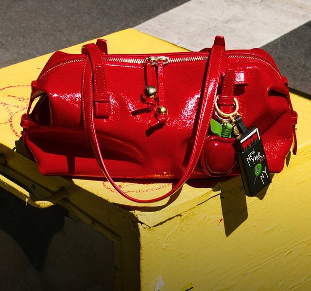 Red patent leather handbag with gold hardware and charm displayed on yellow branded box.