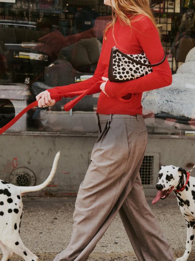 Woman in red sweater and dalmatian-print crossbody bag walking a dalmatian dog on a city street.